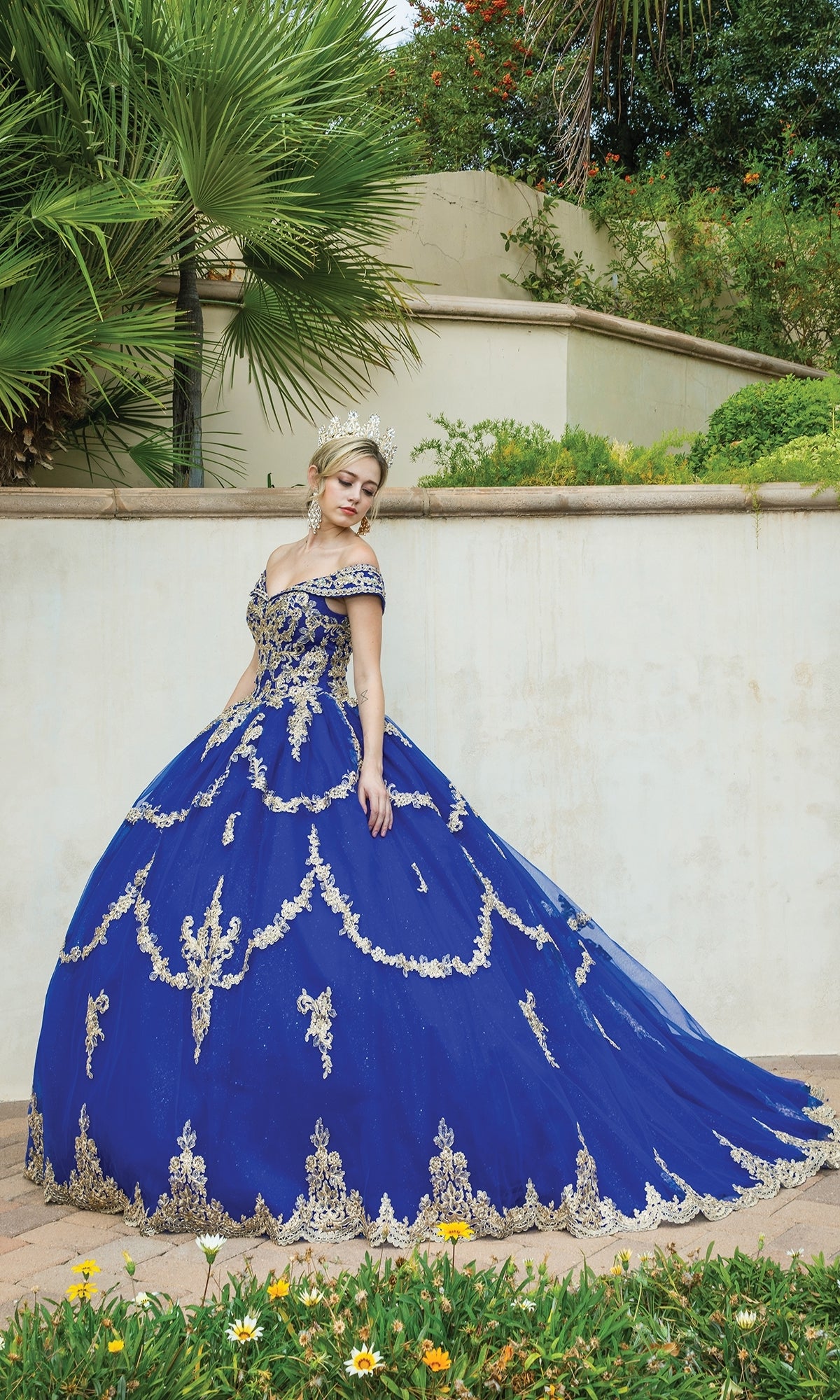A woman stands outdoors in front of a cream wall and greenery, wearing the Dancing Queen Quinceanera Dress 1572—an ornate royal blue ball gown with gold lace embroidery—and a tiara.